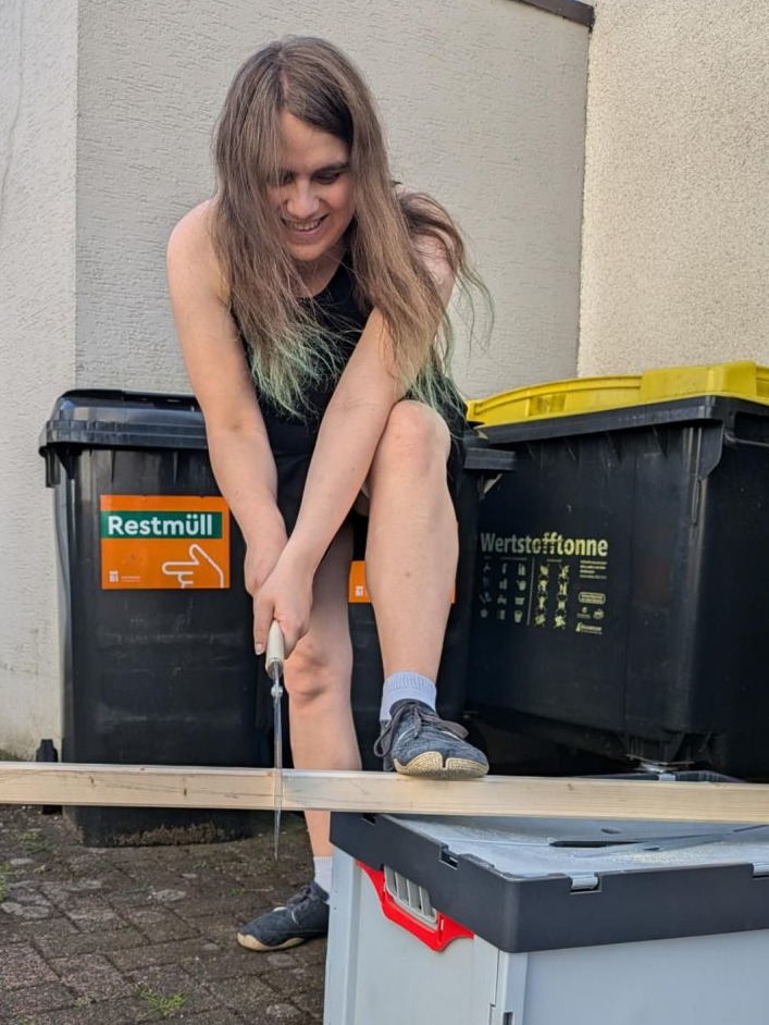 Ein Bild von Zora, wie sie mit einer Zugsäge eine Holzleiste zusägt. Die Holzleiste liegt auf einer grauen Eurobox, Zora hat einen Fuß auf der Leiste um sie dort zu fixieren. Zora schaut konzentriert nach unten, aber lächelt, die Haare fallen ihr ins Gesicht. Sie trägt ein (für ihre Verhältnisse) recht kurzes schwarzes Kleid. Im Hintergrund sind mehrere Mülltonnen.